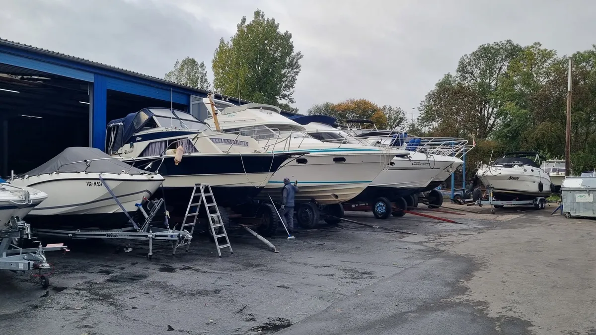 Motorboote und Motoryachten stehen auf Trailern vor einer offenen blauen Halle, ein Mann reinigt einen Rumpf
