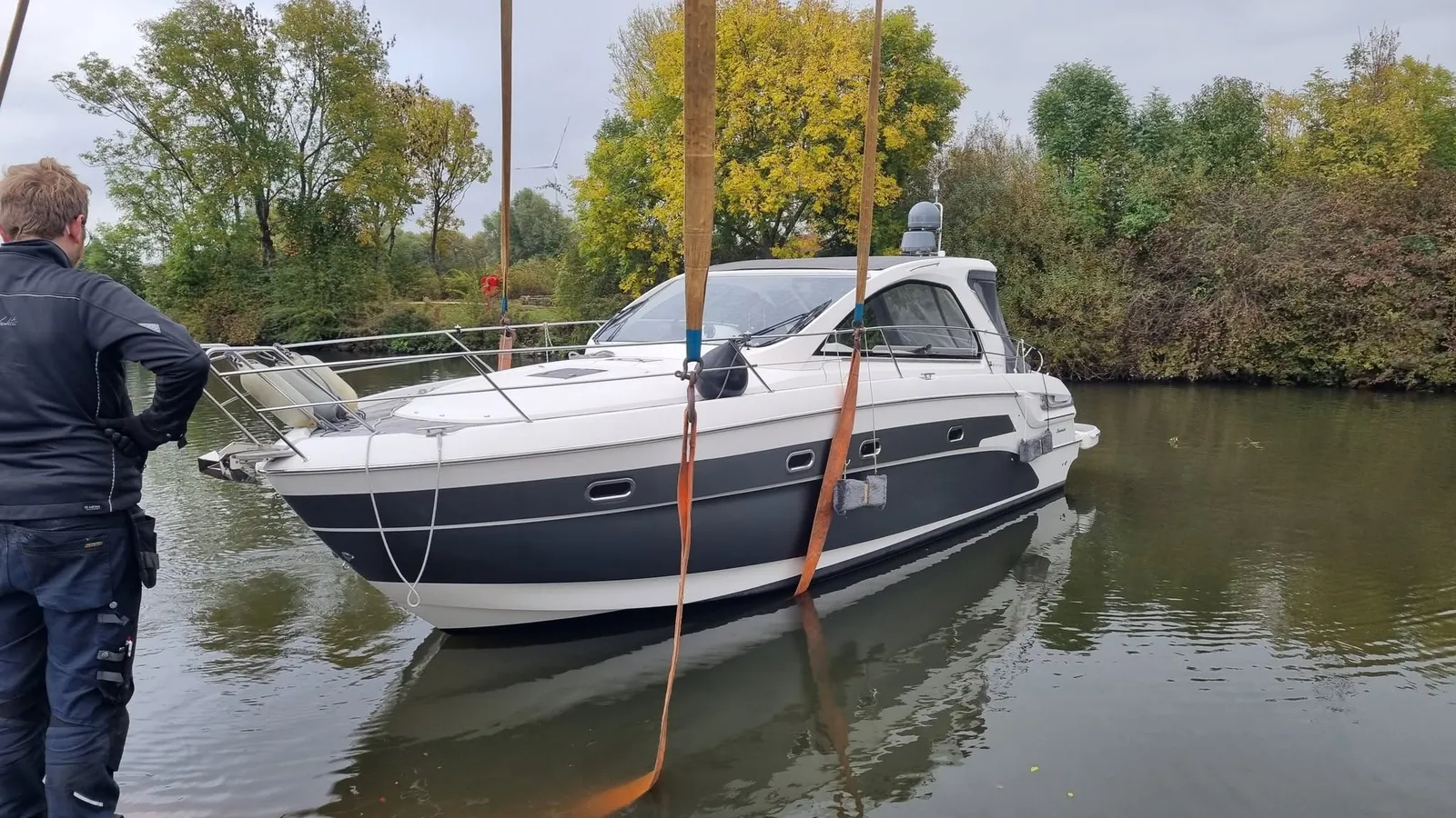Ein Mann in Arbeitskleidung steht am Ufer, vor ihm wird eine Motoryacht in Gurten aus dem Wasser gehoben