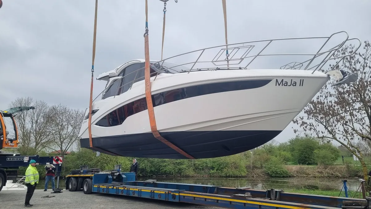 Eine weiße Motoryacht mit großen Rumpffenstern hängt in Gurten am Mobilkran über einem Tieflader, dahinter Bäume am Ufer